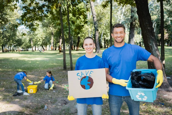 Paar hält Plakat mit Globus, Aufschrift und Müllbehälter in der Hand, während Kinder auf verschwommenem Hintergrund Müll sammeln, Ökologiekonzept — Stockfoto