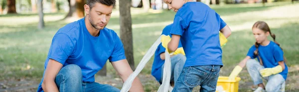 Père et fils ramassant des déchets plastiques dans la forêt près de la mère et de la fille sur fond flou, concept écologique, bannière — Photo de stock