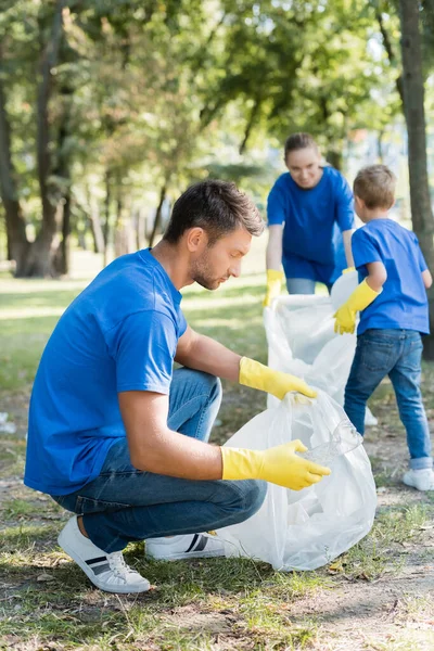 Mann hält recycelte Tüte in der Hand, während Familie Müll auf verschwommenem Hintergrund sammelt, ökologisches Konzept — Stockfoto