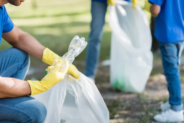 Vista cortada do homem segurando garrafa de plástico e saco reciclado perto da família no fundo turvo, conceito de ecologia — Fotografia de Stock