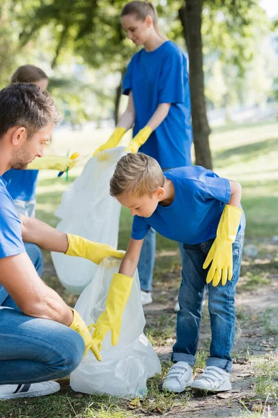 Vater und Sohn sammeln Müll in Recyclingtüte in der Nähe von Mutter und Tochter auf verschwommenem Hintergrund, ökologisches Konzept — Stockfoto