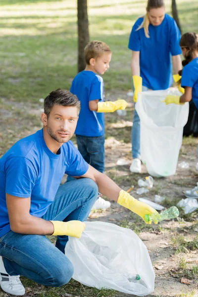 Mann blickt in Kamera, während Familie Plastikmüll auf verschwommenem Hintergrund sammelt, Ökologiekonzept — Stockfoto