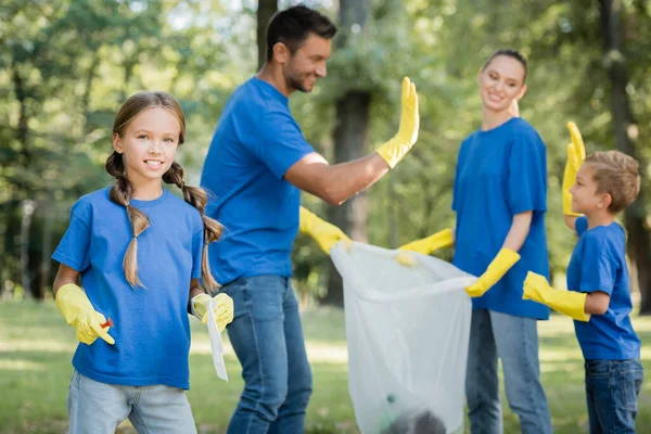 Mädchen blickt in die Kamera, während Eltern eine recycelte Tasche in der Hand halten und auf verschwommenem Hintergrund eine hohe Fünf geben, Ökologiekonzept — Stockfoto