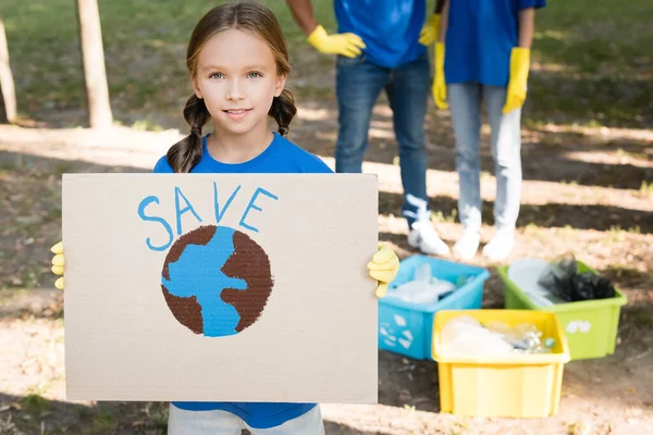 Menina segurando cartaz com globo e salvar inscrição, e os pais perto de recipientes de lixo no fundo borrado, conceito de ecologia — Fotografia de Stock