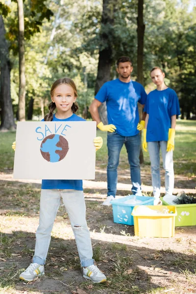 Lächelndes Mädchen mit Plakat mit Globus und Schriftzug, Eltern in der Nähe von Müllcontainern auf verschwommenem Hintergrund, Umweltkonzept — Stockfoto