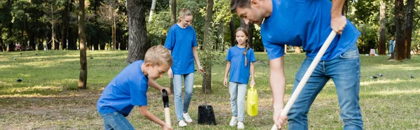 Mother and daughter carrying young tree and watering can near father and son with shovels on blurred foreground, ecology concept, banner — Stock Photo