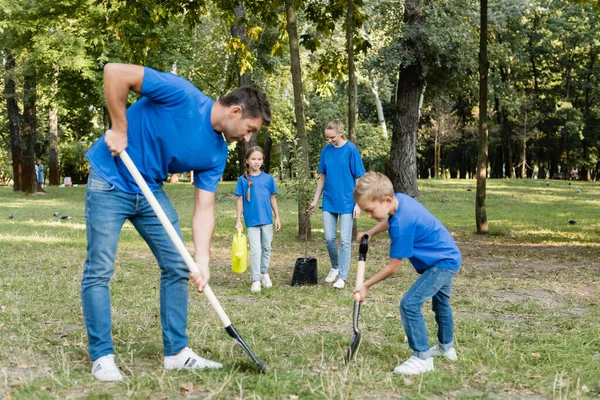 Father and son digging ground while mother and daughter carrying young tree and watering can, ecology concept — Stock Photo