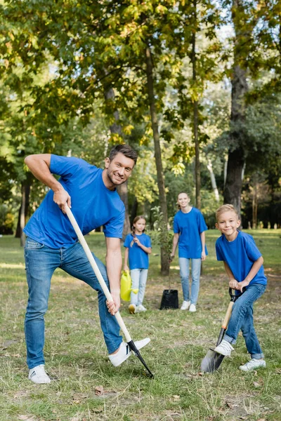 Père et fils souriant à la caméra tout en creusant le sol, et mère avec fille tenant jeune arbre et arrosoir sur fond flou, concept d'écologie — Photo de stock