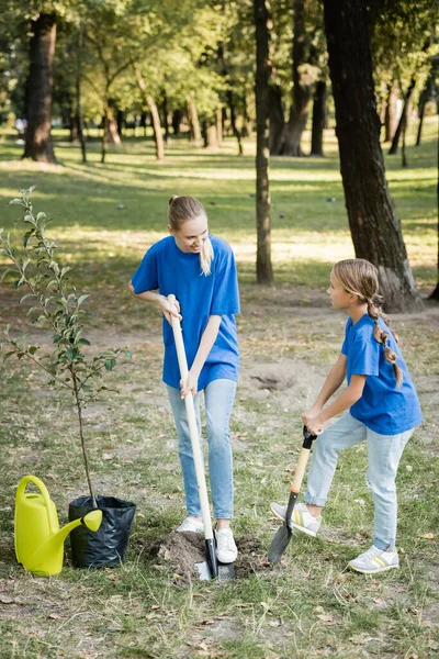 Daughter and mother digging ground with shovels near watering can and young tree, ecology concept — Stock Photo