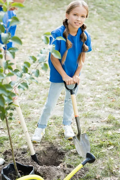Girl looking at camera while holding shovel near mother and young green tree, ecology concept — Stock Photo