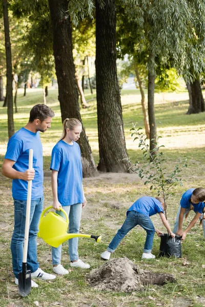Crianças carregando árvore jovem, enquanto os pais de pé com pá e regador pode, conceito de ecologia — Fotografia de Stock
