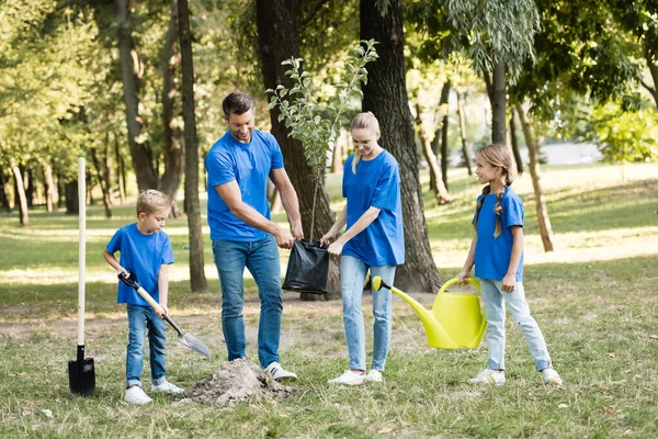 Garçon creuser terrain, fille tenant la boîte d'eau, et les parents portant jeune arbre, concept d'écologie — Photo de stock