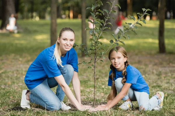 Mère et fille souriantes regardant la caméra tout en plantant un jeune arbre dans le parc, concept d'écologie — Photo de stock