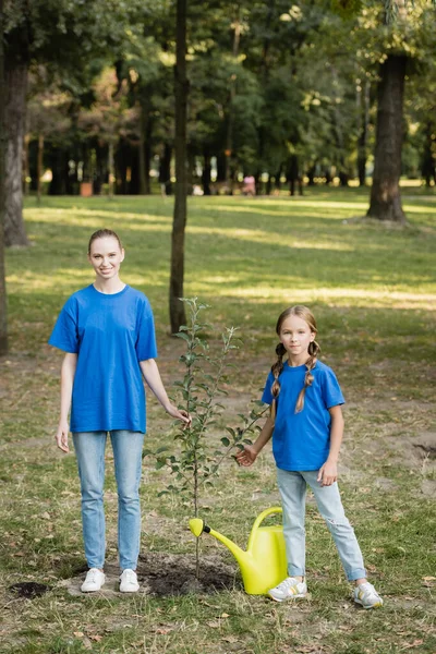 Happy mother and daughter looking at camera while standing near young planted tree, ecology concept — Stock Photo
