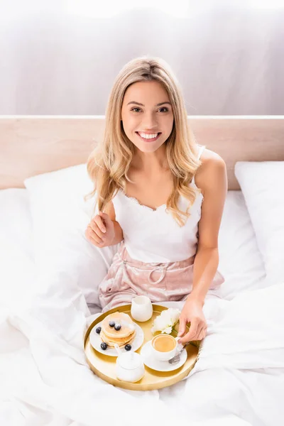 Mujer alegre mirando a la cámara mientras sostiene la taza de café cerca de panqueques y flores en el desayuno trey en la cama - foto de stock