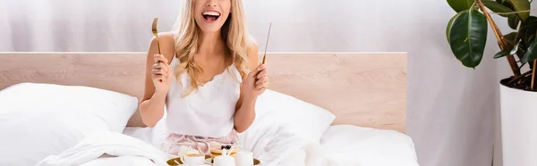 Cropped view of cheerful woman holding cutlery near breakfast on tray on bed, banner — Stock Photo