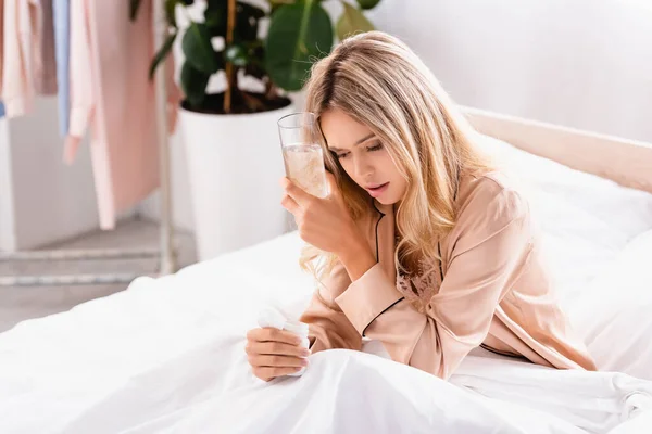 Женщина в пижаме держит стакан воды и банку с таблетками на кровати — стоковое фото