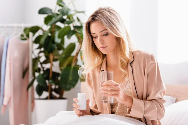 Young woman looking at jar with pills and holding glass of water on bed — Stock Photo