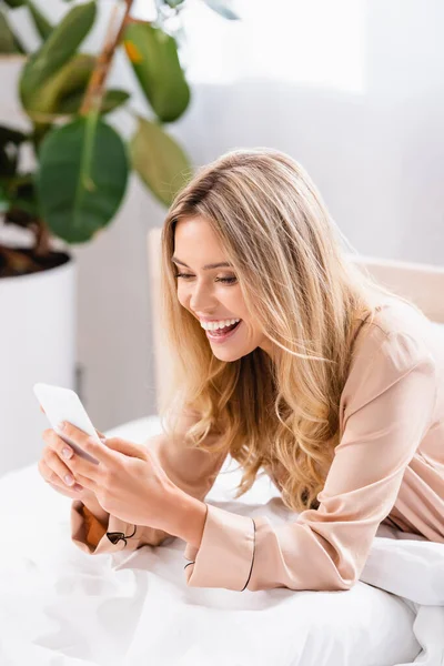 Donna sorridente in pigiama con smartphone sul letto — Foto stock