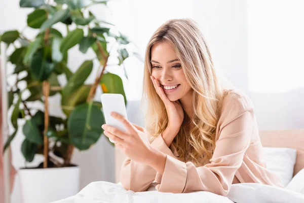 Jeune femme avec la main près de la joue en utilisant smartphone sur le lit le matin — Photo de stock