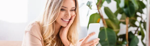 Улыбающаяся женщина с помощью смартфона на размытом переднем плане дома, баннер — стоковое фото