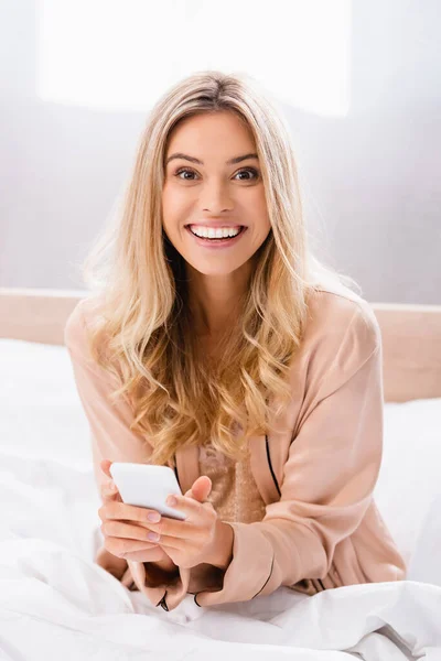 Young woman in pajamas smiling at camera while using smartphone on bed — Stock Photo