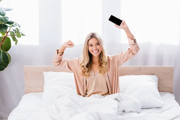 Весела жінка в піжамі тримає смартфон з порожнім екраном на ліжку вранці — стокове фото