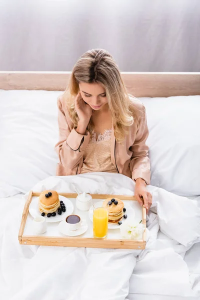Young woman holding tray with pancakes, coffee and orange juice on bed — Stock Photo