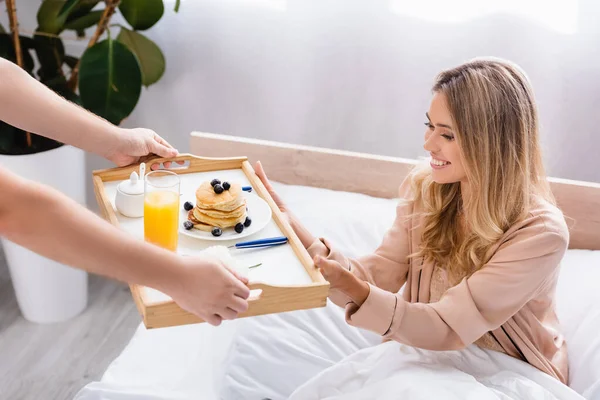 Man giving breakfast tray with pancakes and orange juice near smiling girlfriend on bed — Stock Photo