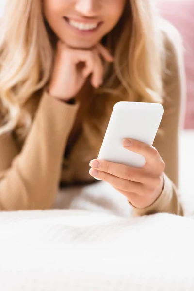 Vista cortada do smartphone na mão de mulher sorridente no fundo embaçado na cama — Fotografia de Stock