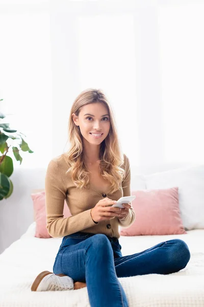 Smiling woman in jeans using smartphone while sitting on bed at home — Stock Photo