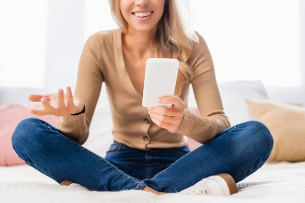 Smartphone en la mano de una mujer sonriente sentada en la cama sobre un fondo borroso - foto de stock