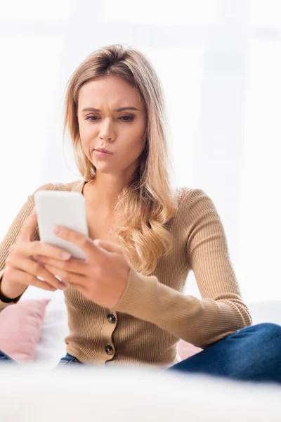 Pensive woman using smartphone on blurred foreground while sitting on bed — Stock Photo