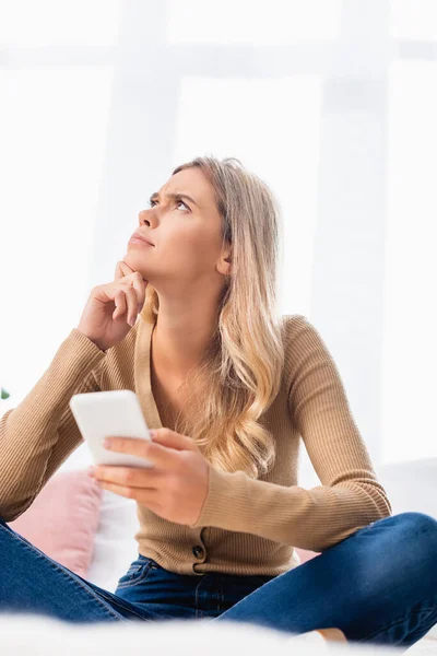 Thoughtful woman looking away while holding smartphone on blurred foreground on bed — Stock Photo