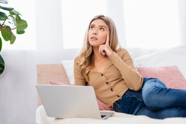 Freelancer pensativo mirando hacia otro lado cerca del ordenador portátil en la cama - foto de stock