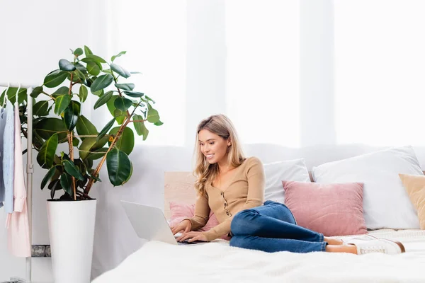 Sourire freelance en utilisant un ordinateur portable tout en travaillant sur le lit à la maison — Photo de stock