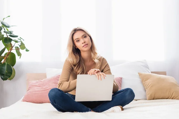 Jovem mulher olhando para a câmera enquanto segurando laptop no quarto — Fotografia de Stock