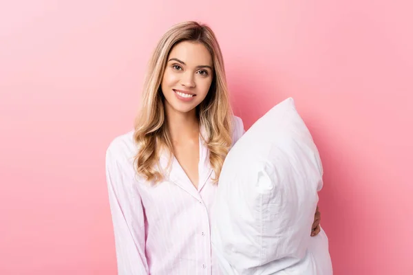 Young woman in pajamas smiling at camera and holding pillow on pink background — Stock Photo