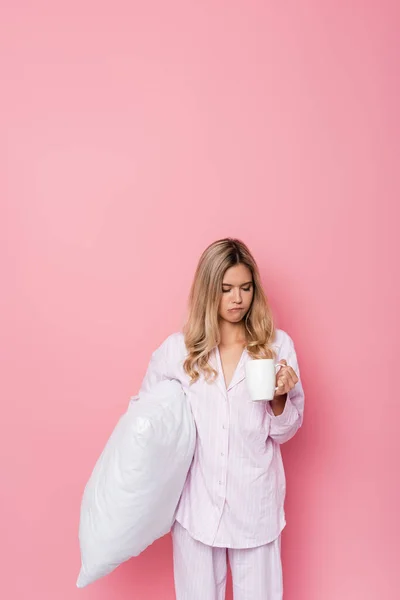 Pensive woman in pajamas holding pillow and cup on pink background — Stock Photo