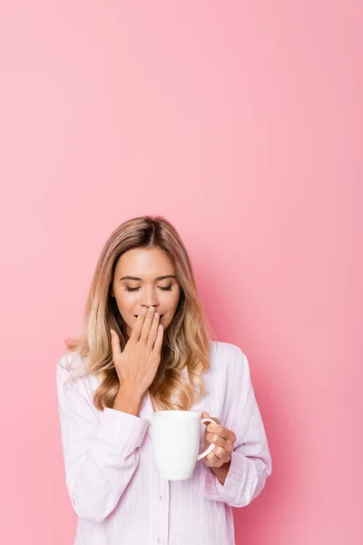 Young woman in pajamas yawning and holding cup on pink background — Stock Photo