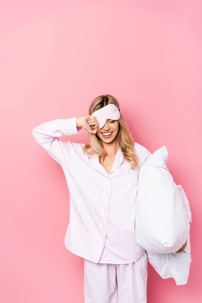 Cheerful woman in blindfold and pajamas holding pillow on pink background — Stock Photo