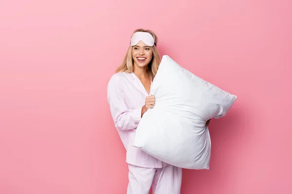 Young woman in pajamas and blindfold holding white pillow on pink background — Stock Photo