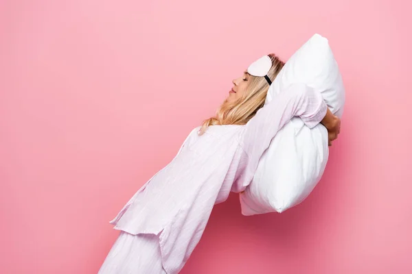 Young woman in pajamas and blindfold holding pillow near head on pink background — Stock Photo