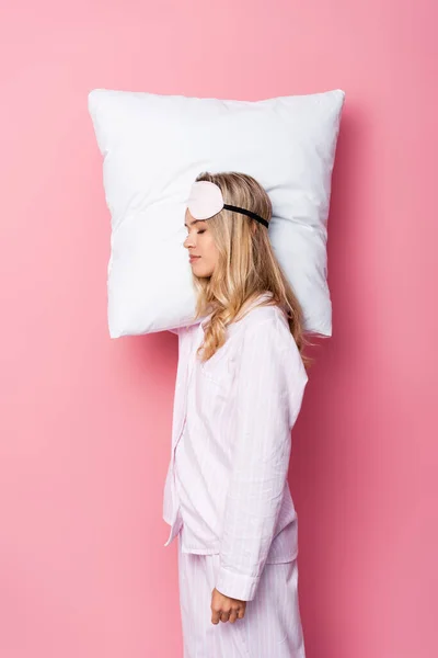 Young woman in pajamas and blindfold holding pillow on pink background — Stock Photo