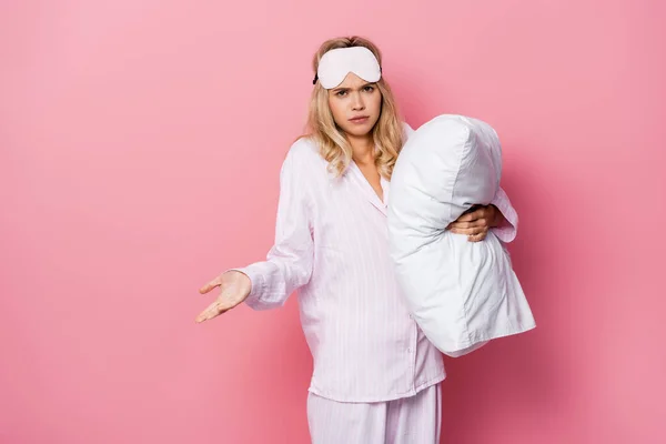 Femme confus dans les yeux bandeau et pyjama tenant oreiller et montrant geste haussant les épaules sur fond rose — Photo de stock