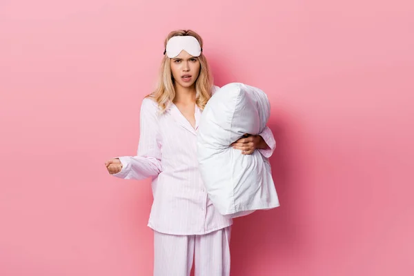 Mujer enojada en pijama y con los ojos vendados sosteniendo almohada sobre fondo rosa - foto de stock