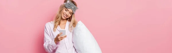 Mujer sonriente con los ojos vendados y pijamas sosteniendo la almohada y usando un teléfono inteligente sobre fondo rosa, pancarta - foto de stock