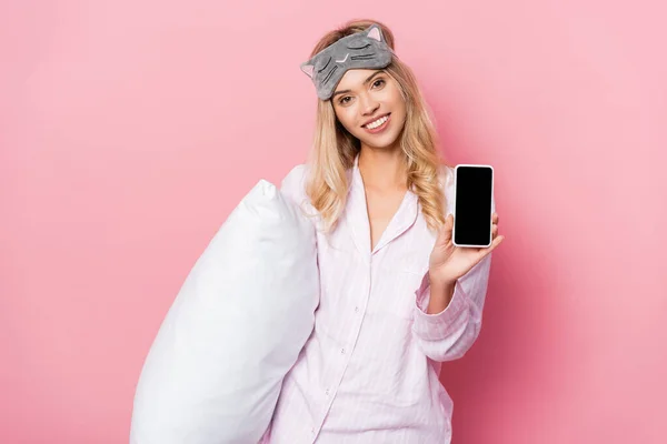Mujer alegre en venda y pijama sosteniendo almohada y mostrando teléfono inteligente con pantalla en blanco sobre fondo rosa - foto de stock