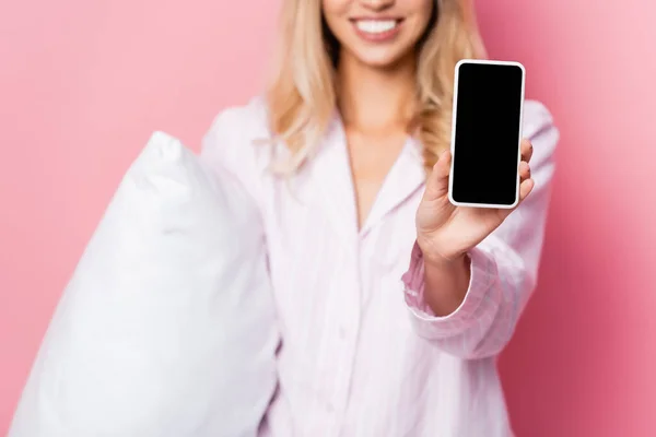 Vista recortada del smartphone con pantalla en blanco en la mano de una mujer sonriente en pijama con almohada borrosa sobre fondo rosa - foto de stock