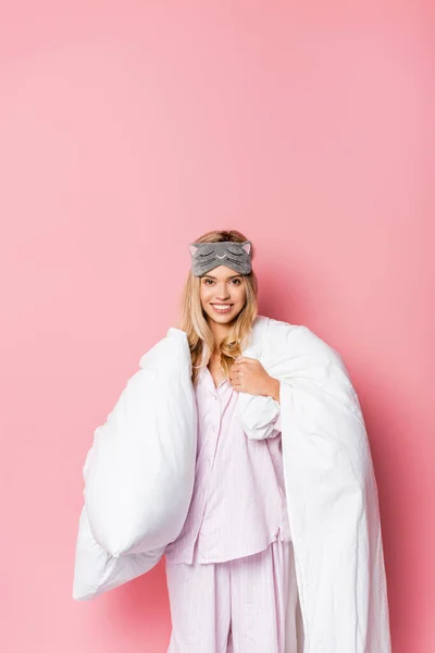 Cheerful woman in pajamas and blindfold holding blanket on pink background — Stock Photo